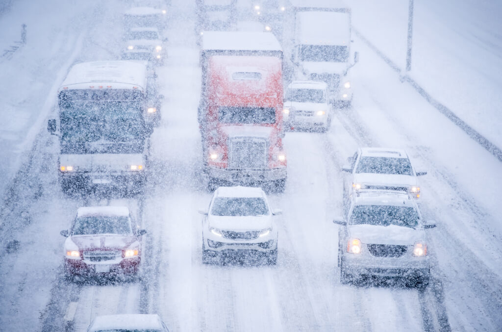 Photograph of several cars, a bus and a tractor trailer driving down a snow covered highway.